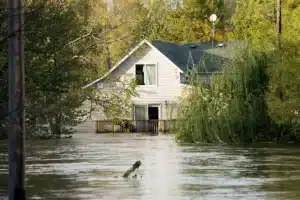 Suburban house surrounded by floodwater, with water reaching the deck and trees partially submerged.