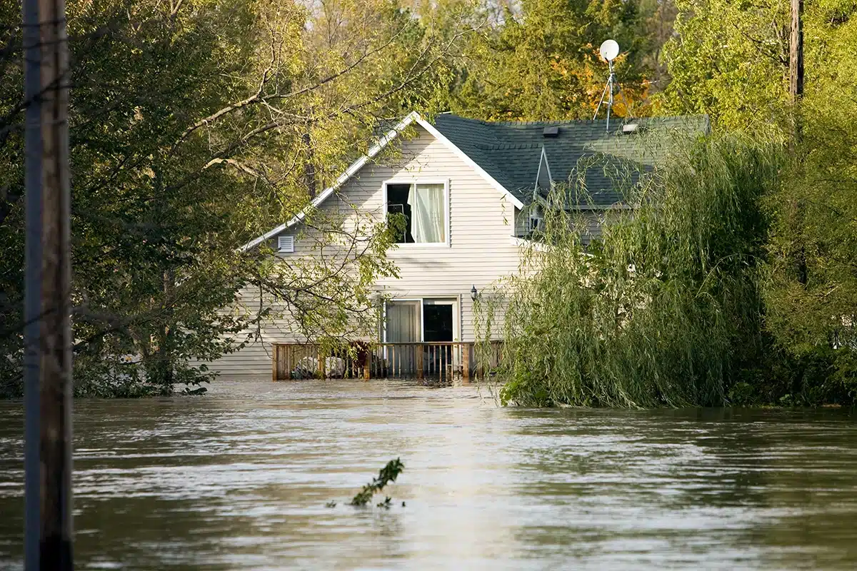 Suburban house surrounded by floodwater, with water reaching the deck and trees partially submerged.